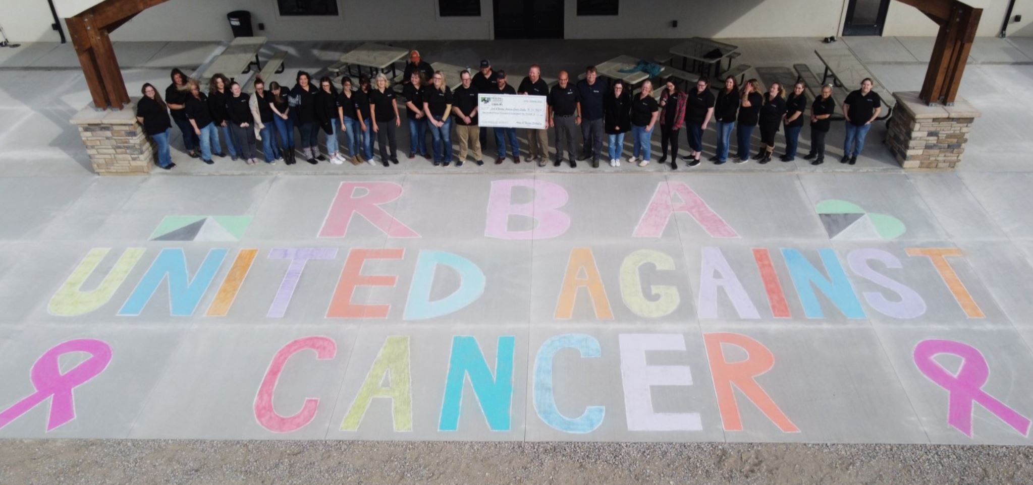 RbA United Against Cancer written on chalk in front of the team from Wyoming. They are holding a large check for a donation.